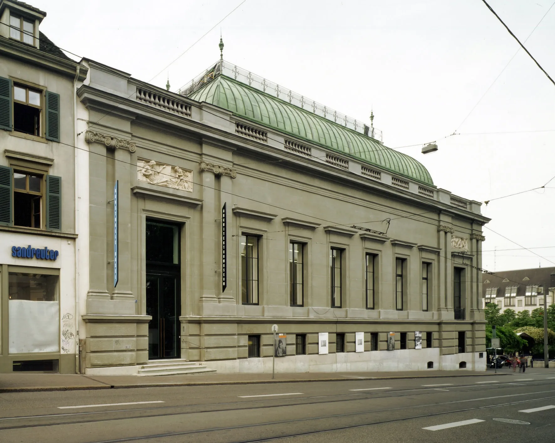 Vista a nivel de calle de la fachada del edificio de la Kunsthalle Basel, que presenta un distintivo techo de cúpula de cobre verde, arquitectura de piedra clásica con columnas y grandes ventanas rectangulares, con líneas de tranvía en la calle frente al edificio.