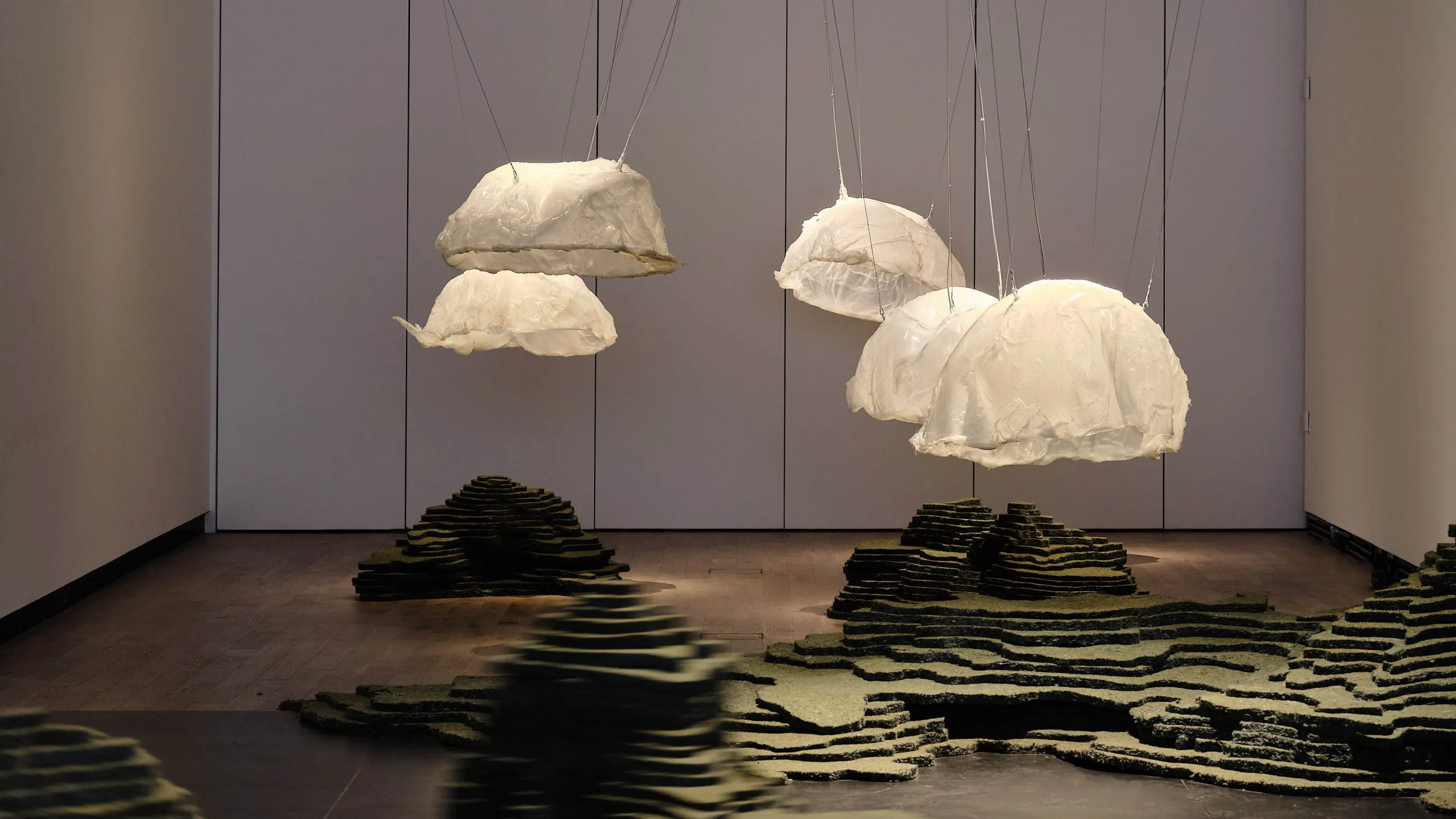 Suspended white cloud-like sculptures and layered dark sculptural forms on the floor in a gallery