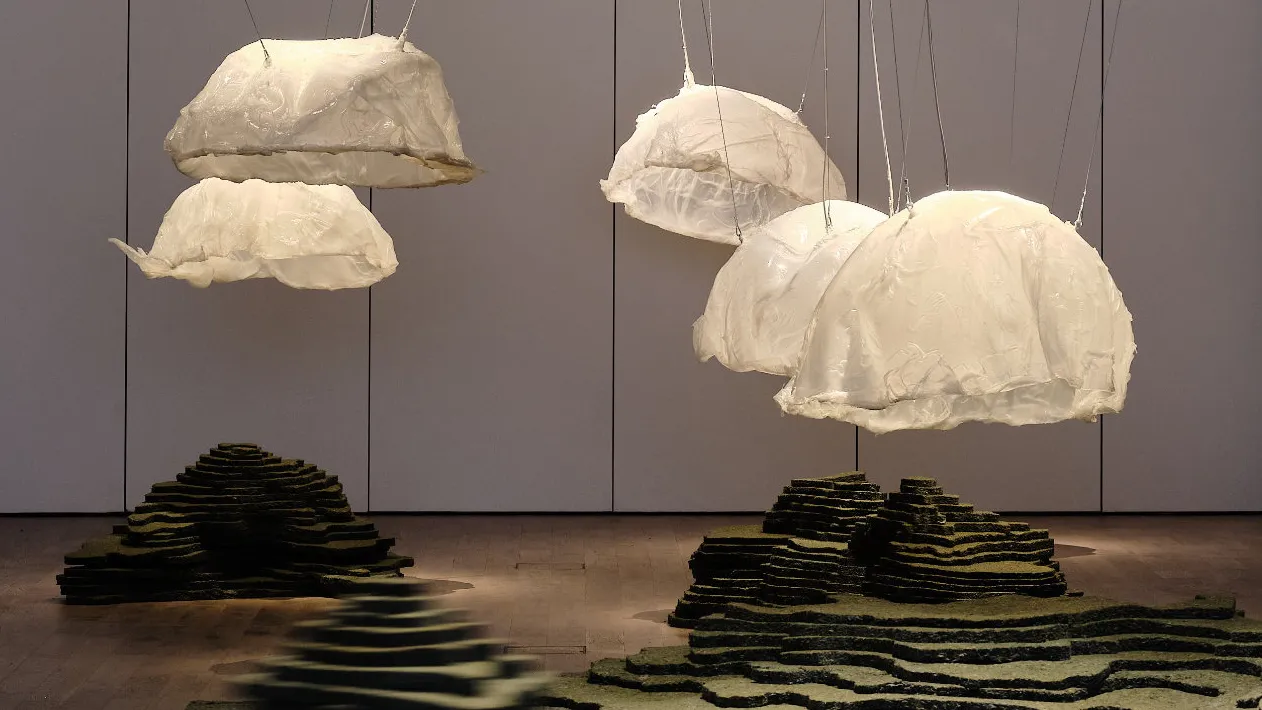 Suspended white cloud-like sculptures and dark layered floor installations in a gallery