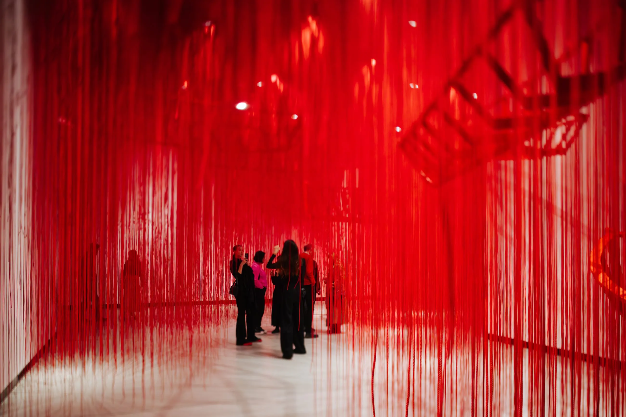 People standing in a gallery space filled with hanging red threads creating a immersive art installation