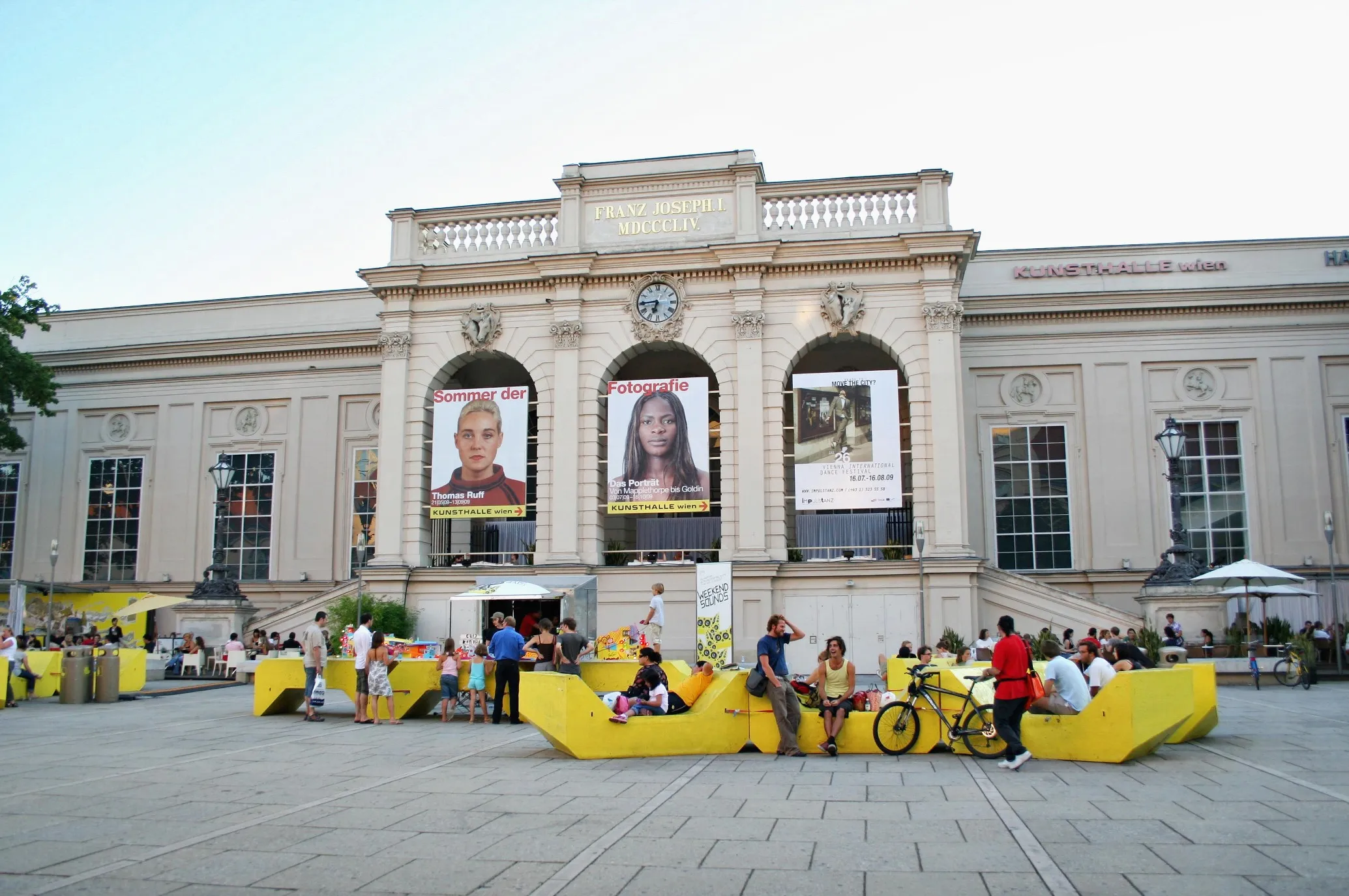 Classical building facade with arched windows and large banners displaying portraits, surrounded by yellow seating and people in a plaza.