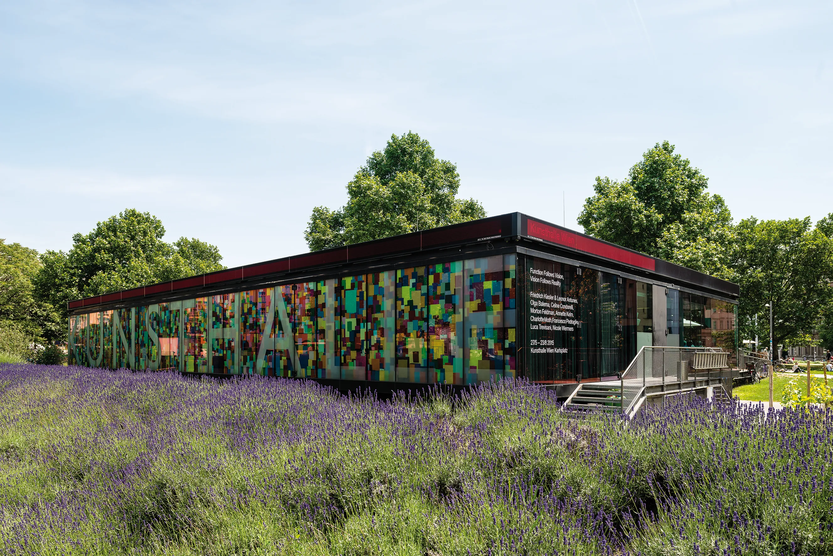 Modern building with colorful pixelated facade and 'KUNSTHALLE' text, surrounded by purple lavender fields and green trees.