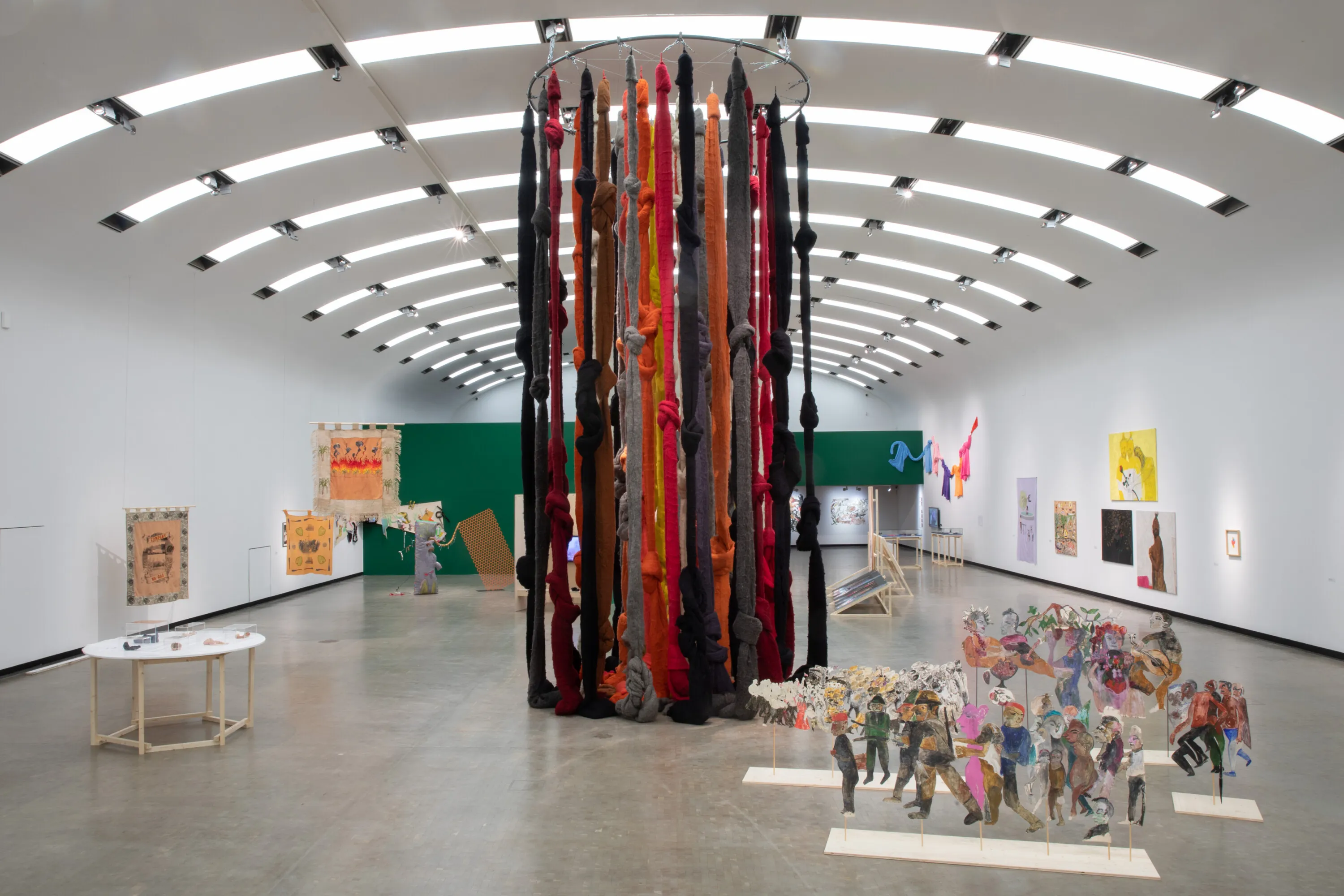 Large cylindrical sculpture made of colorful fabric strips in a modern art gallery with artworks on the walls and floor.