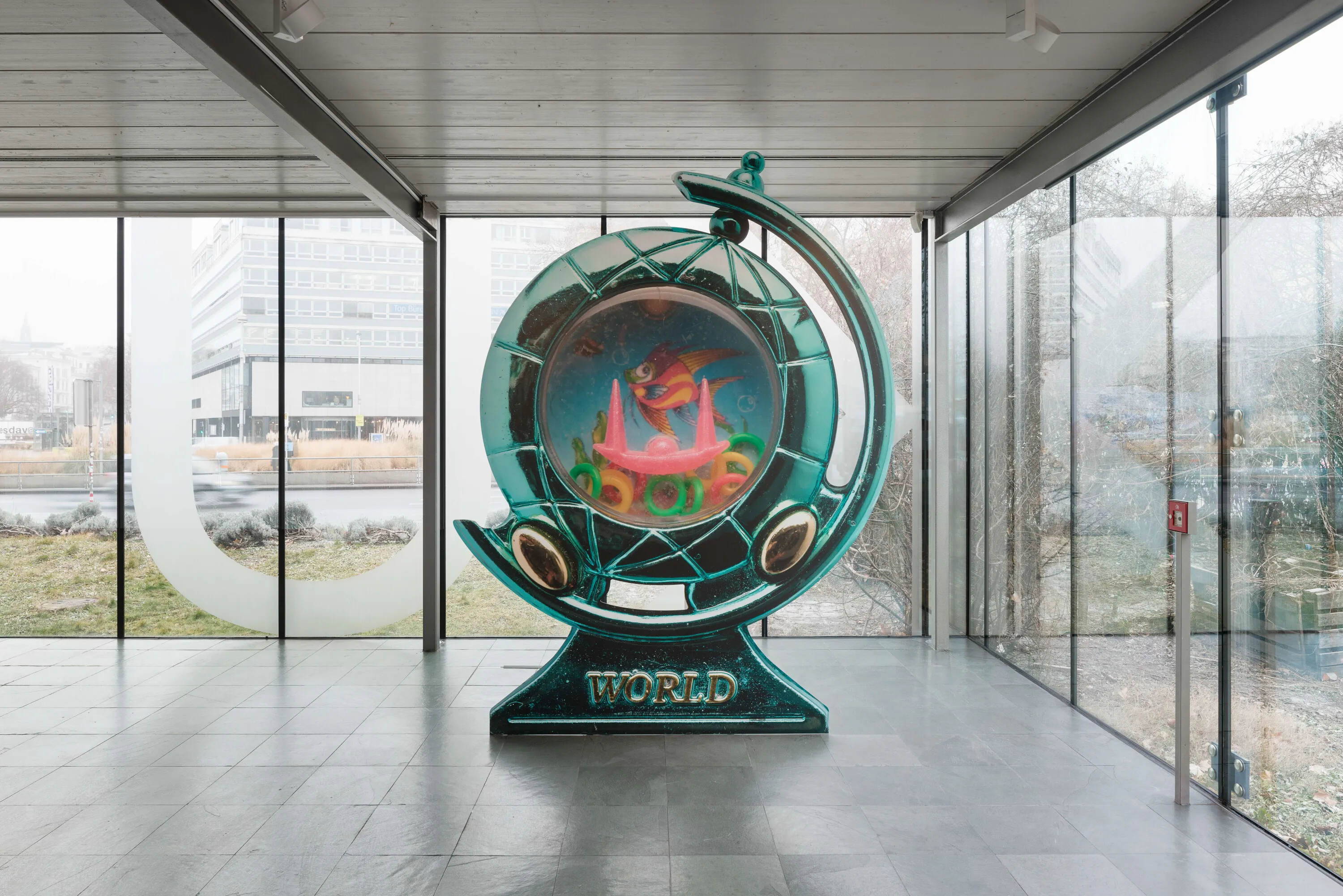 A large, circular sculpture with the word 'WORLD' on its base, featuring colorful internal elements, displayed in a modern gallery with large windows.