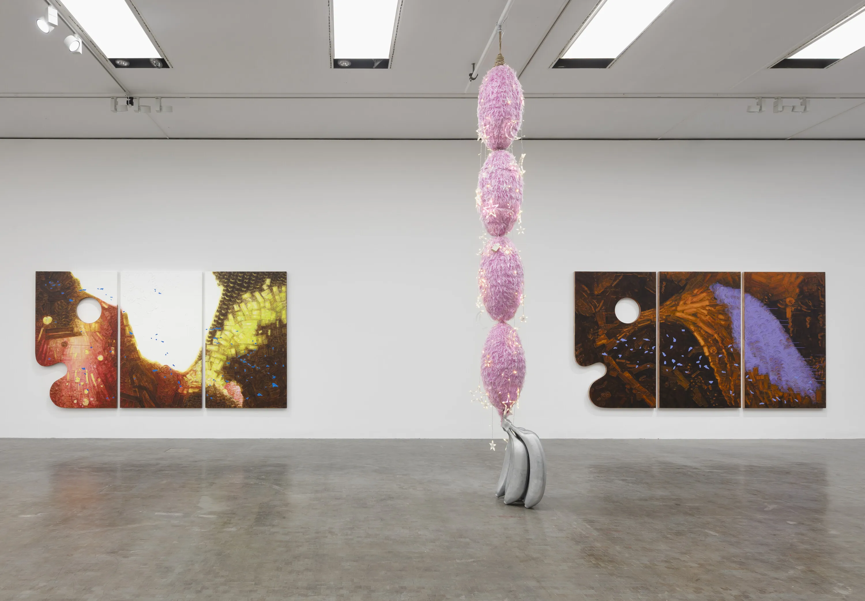 A contemporary art installation with a hanging pink sculpture and two triptych paintings on white walls in a gallery.