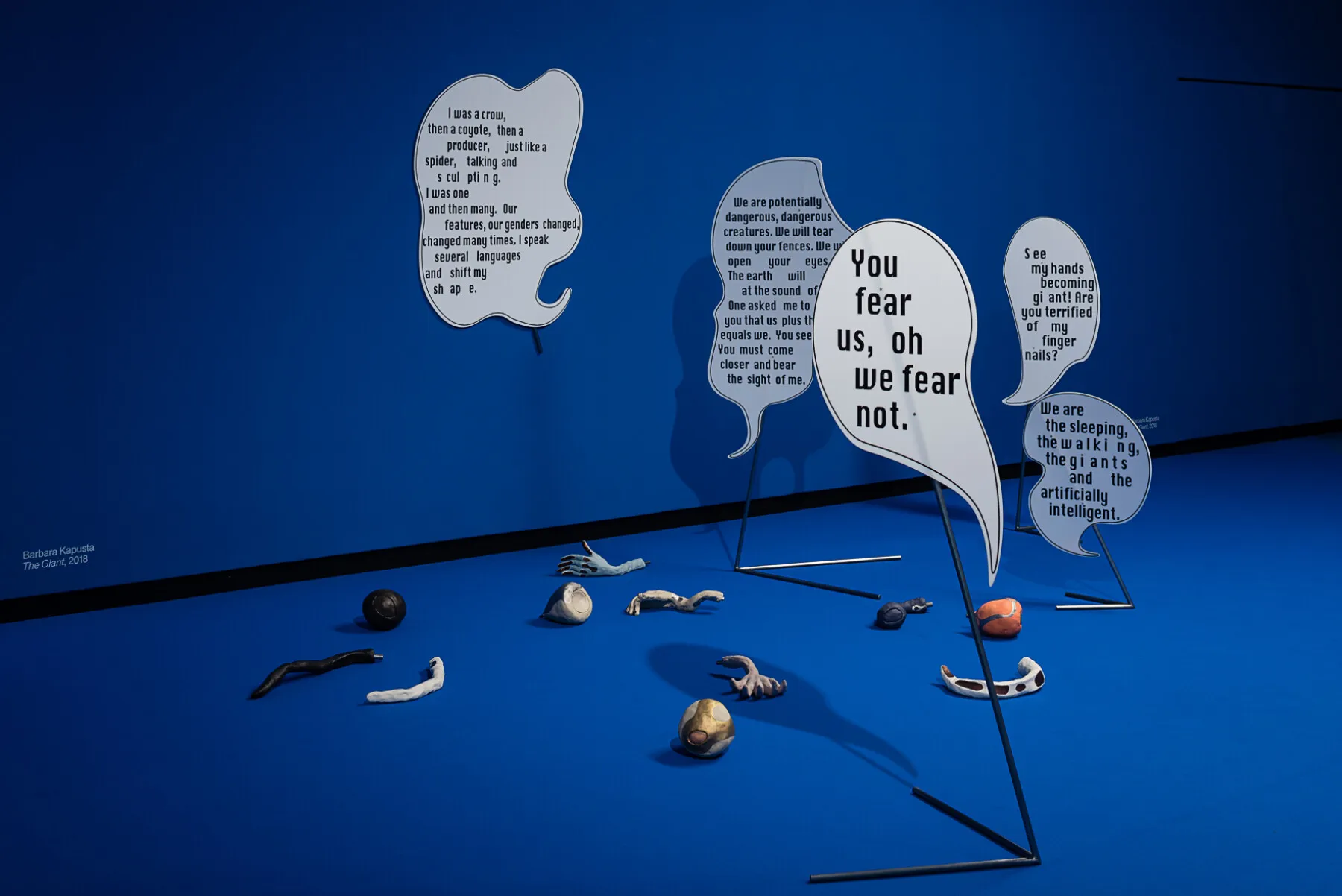 Blue gallery space with speech bubble sculptures containing text and scattered organic objects on the floor
