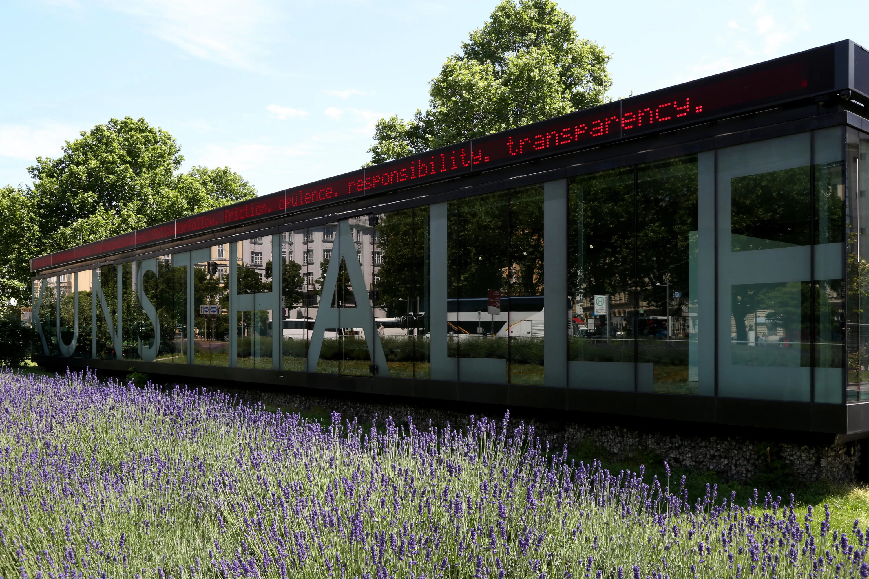 Modern glass building with 'KUNSTHALLE' in large letters and a digital sign, surrounded by lavender flowers