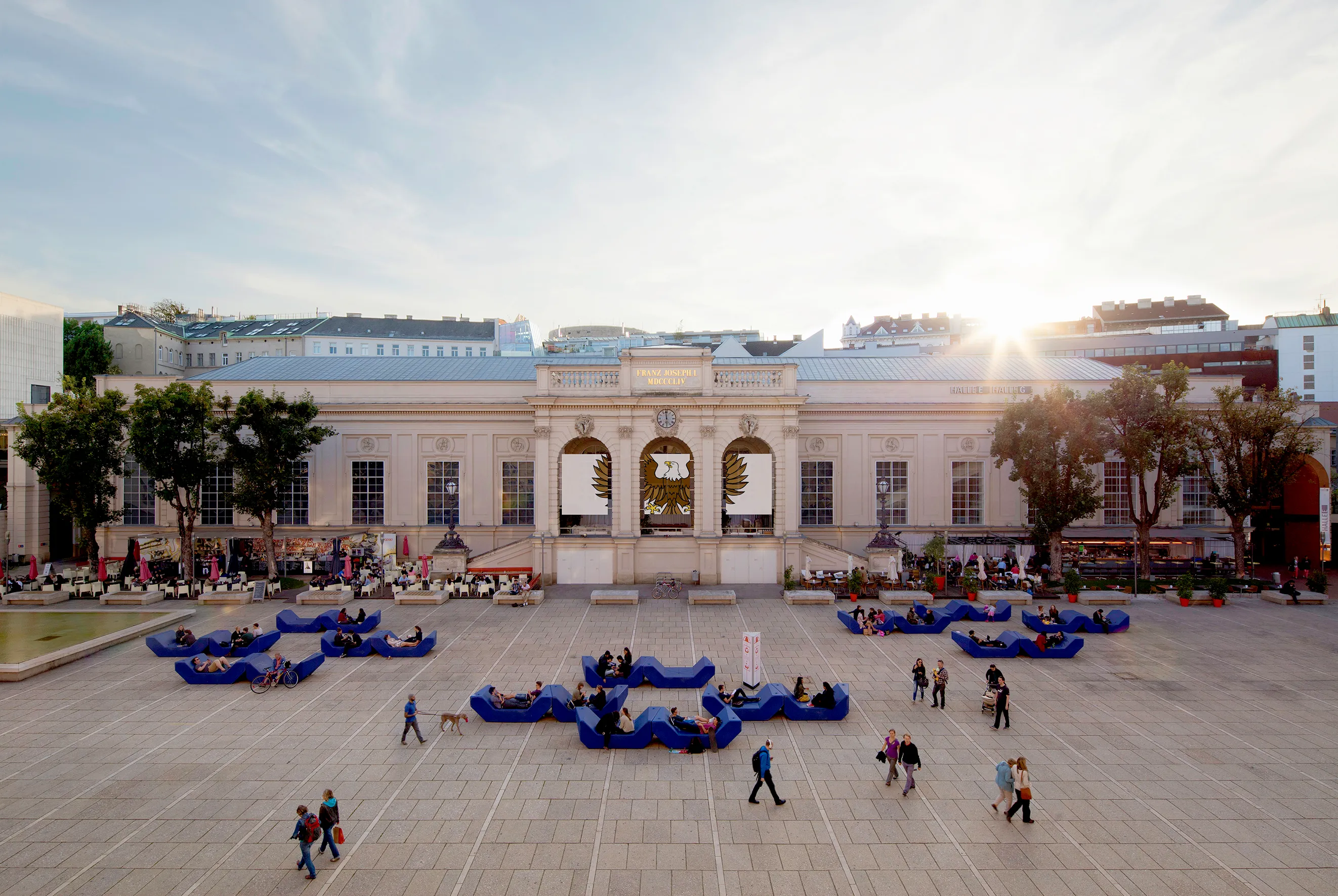 The Kunsthalle Wien building with a large open plaza in front, featuring blue modular seating and people walking, with the sun setting behind the structure.