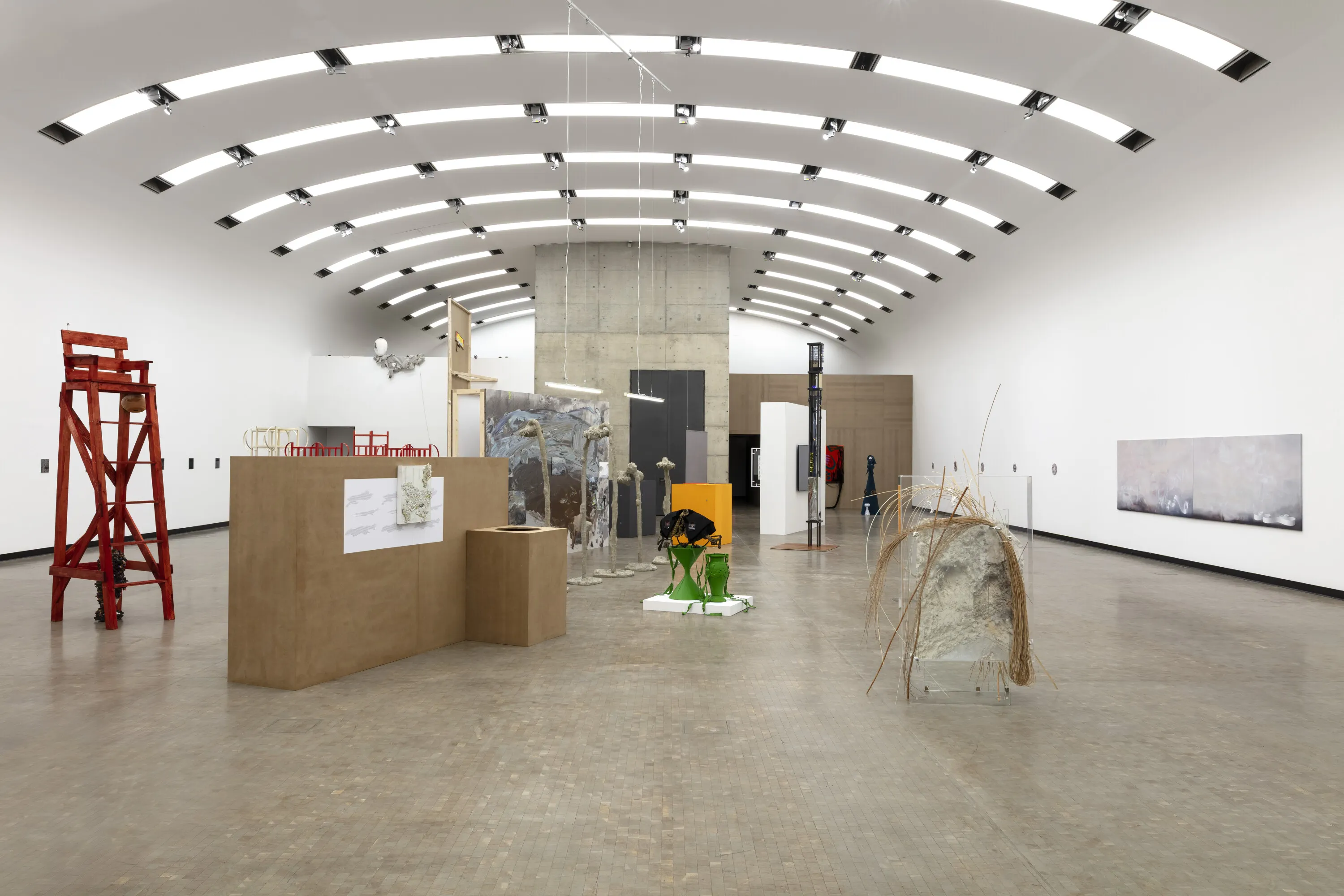 Large gallery space with curved ceiling, white walls, and contemporary art installations including a red ladder, cardboard structures, and a natural material sculpture.