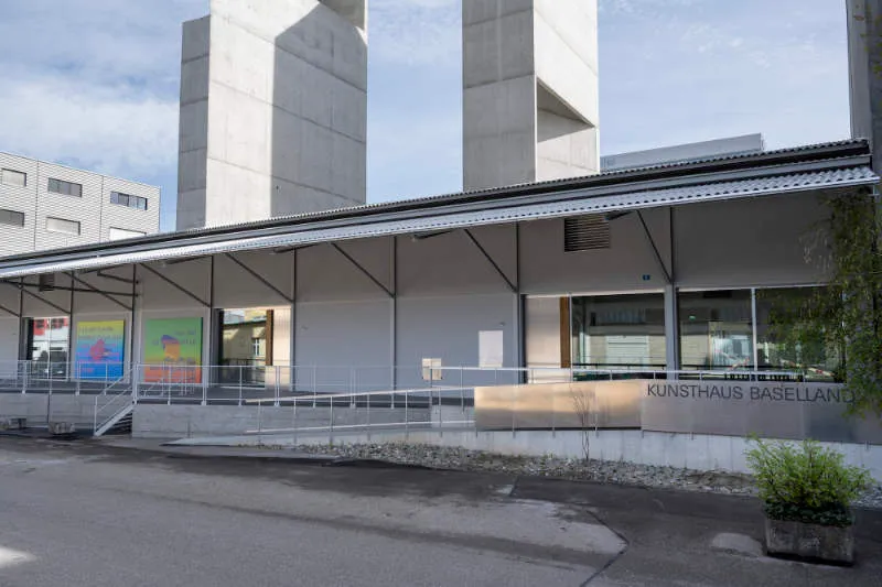 Vista exterior del Kunsthaus Baselland que muestra la fachada del edificio con la rampa de entrada, grandes ventanales y dos altas torres de hormigón al fondo