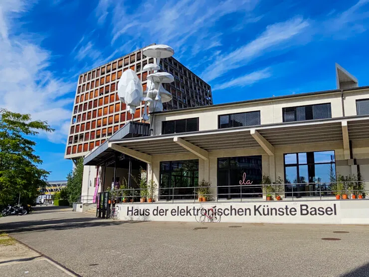 Exterior de edificio moderno con el letrero 'Haus der elektronischen Künste Basel' en un muro bajo, instalación escultórica blanca en la terraza, cielo azul y nubes sobre él