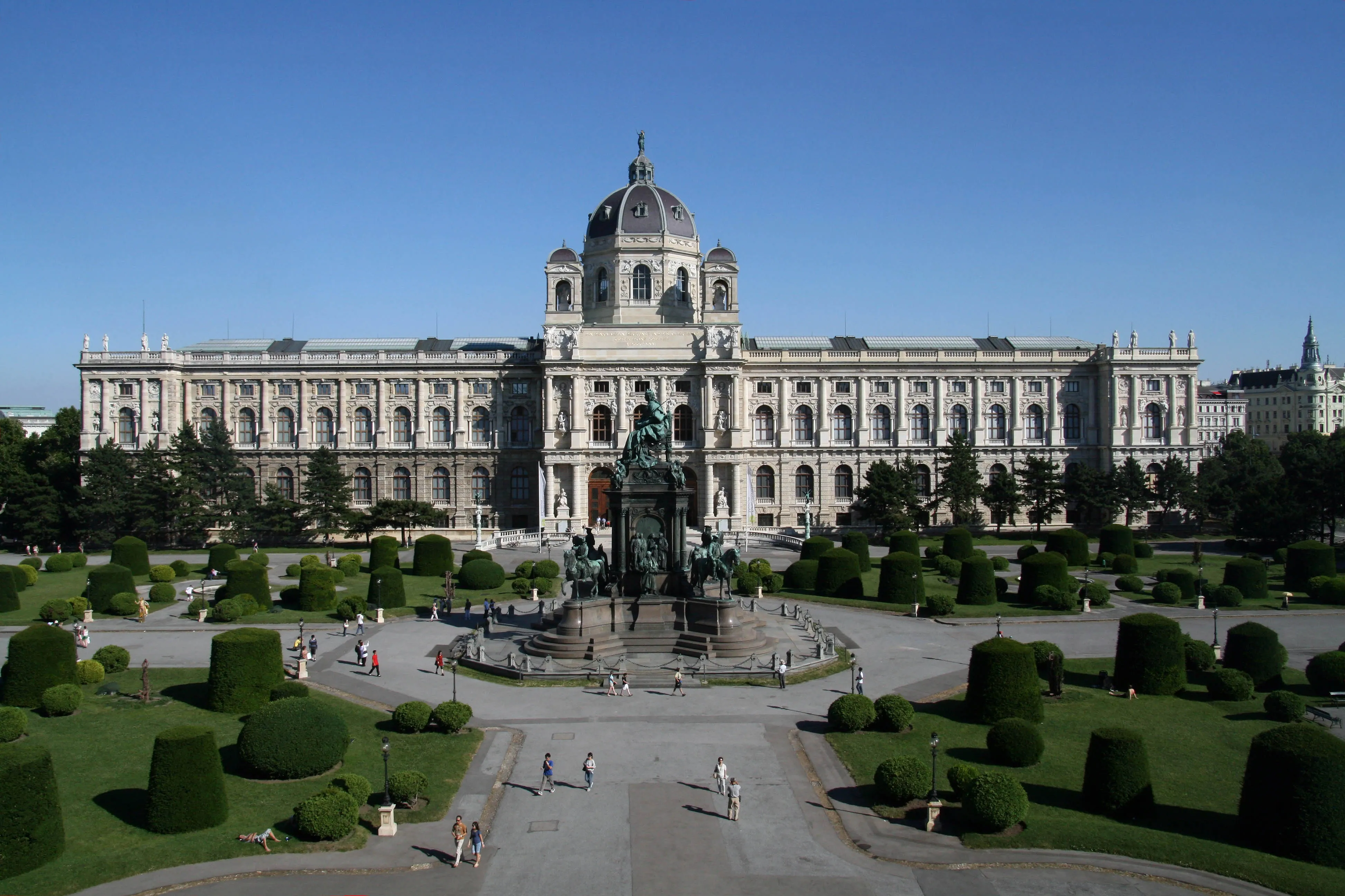 El Kunsthistorisches Museum de Viena, un gran edificio neoclásico con una cúpula central y alas simétricas, visto de frente con un jardín formal y una estatua en primer plano bajo un cielo azul despejado.