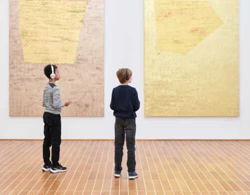 Dos niños pequeños con auriculares están de pie sobre un suelo de parqué de madera, mirando hacia dos grandes pinturas abstractas en tonos dorados y beige en un espacio de galería luminoso.