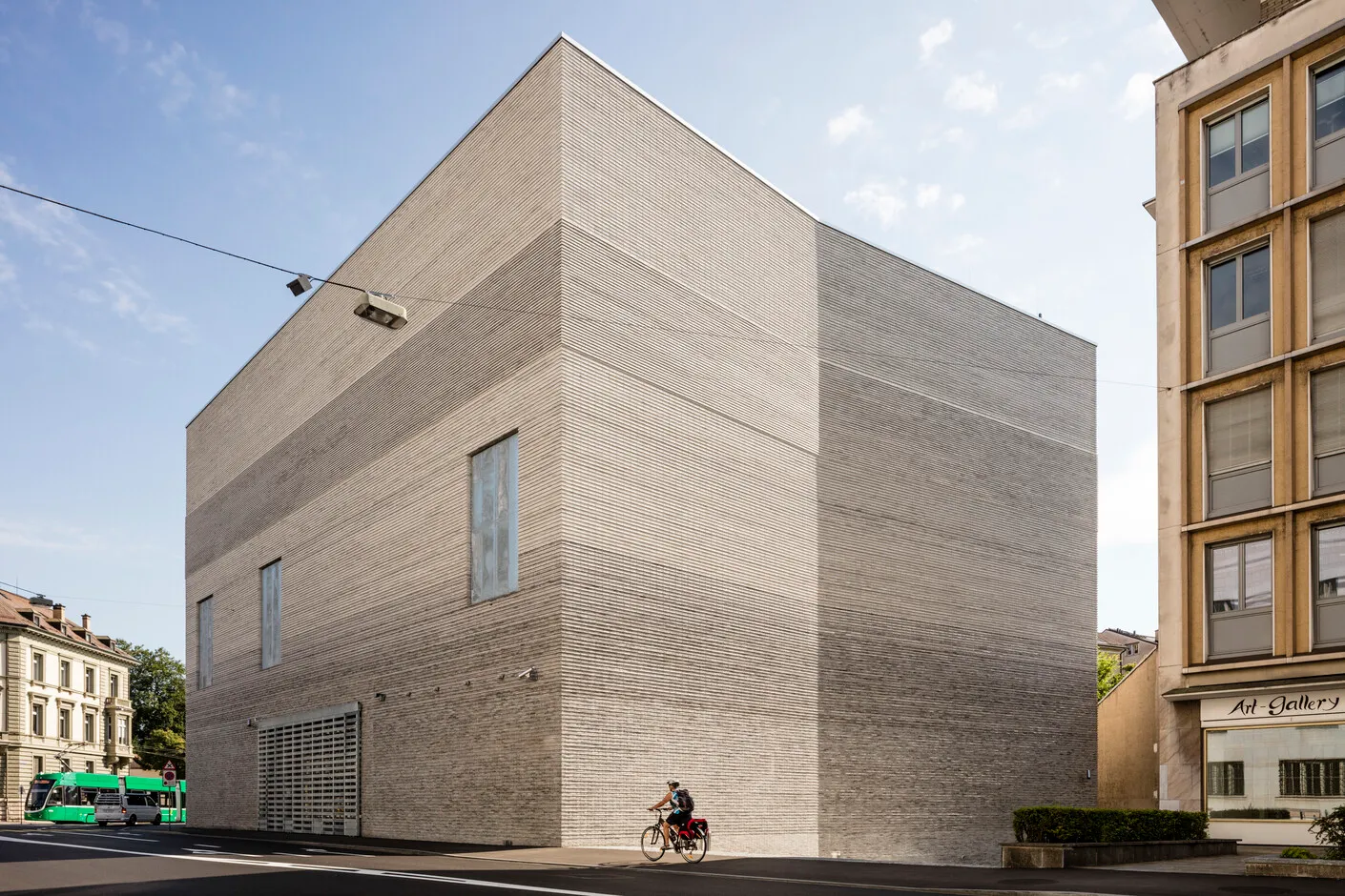 Gran edificio rectangular con fachada de piedra de color gris claro texturizada, ventanas verticales estrechas y un ciclista pasando por la calle.