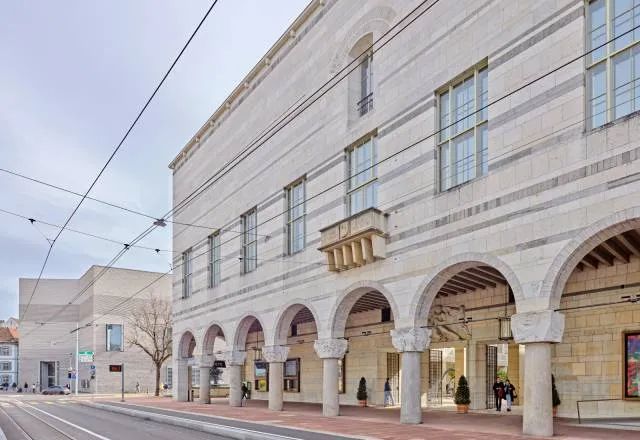 Edificio de piedra del museo con columnata arqueada a lo largo de la fachada que da a la calle, ventanas altas en los pisos superiores y líneas de tranvía elevadas