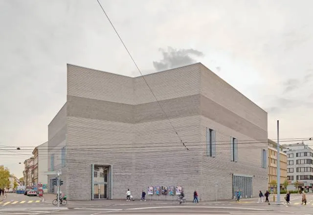 Vista exterior de un edificio grande, moderno y de tonos claros con una característica fachada escalonada, con peatones y líneas de tranvía a nivel de calle.