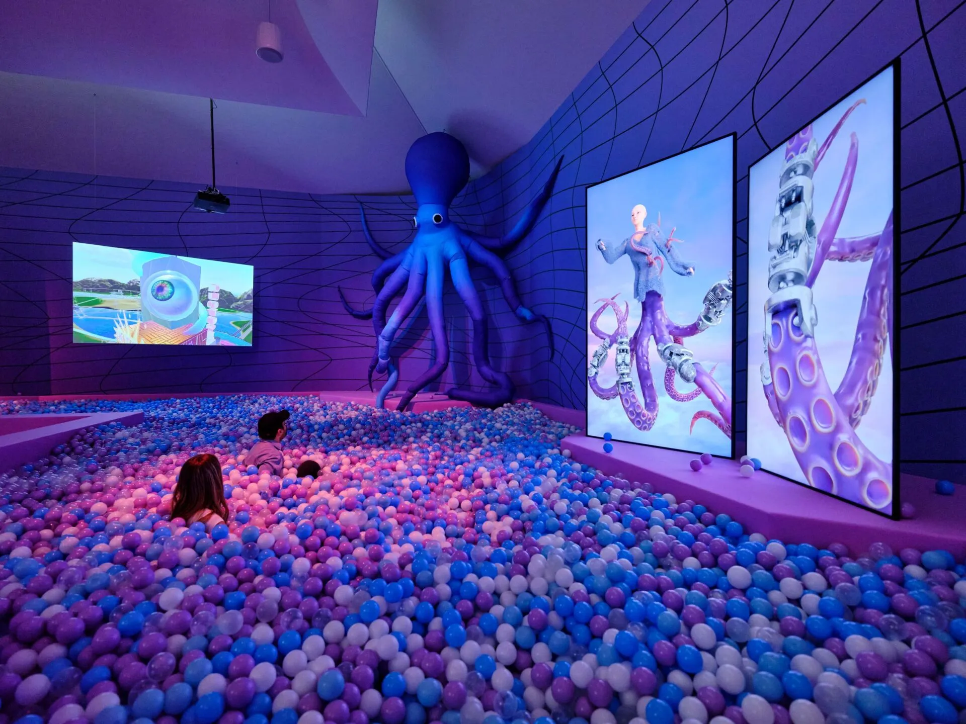 Instalación de arte inmersivo que presenta una gran escultura de pulpo morado, una piscina de bolas llena de bolas azules y rosas, y grandes pantallas de vídeo que muestran imágenes animadas de pulpos en una galería.