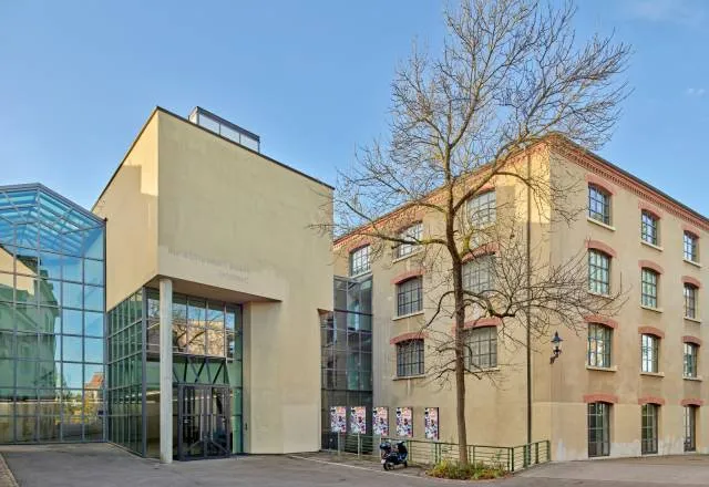 Vista exterior del Kunstmuseum Basel Gegenwart que muestra un moderno pabellón de entrada de cristal conectado a un histórico edificio industrial de color beige con un árbol desnudo en el patio bajo un cielo azul.