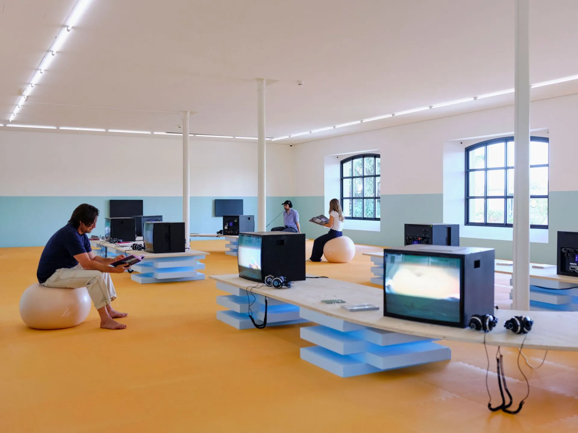 Interior de la galería con monitores de televisión CRT dispuestos sobre plataformas escalonadas de color azul; visitantes sentados en taburetes esféricos observando las pantallas