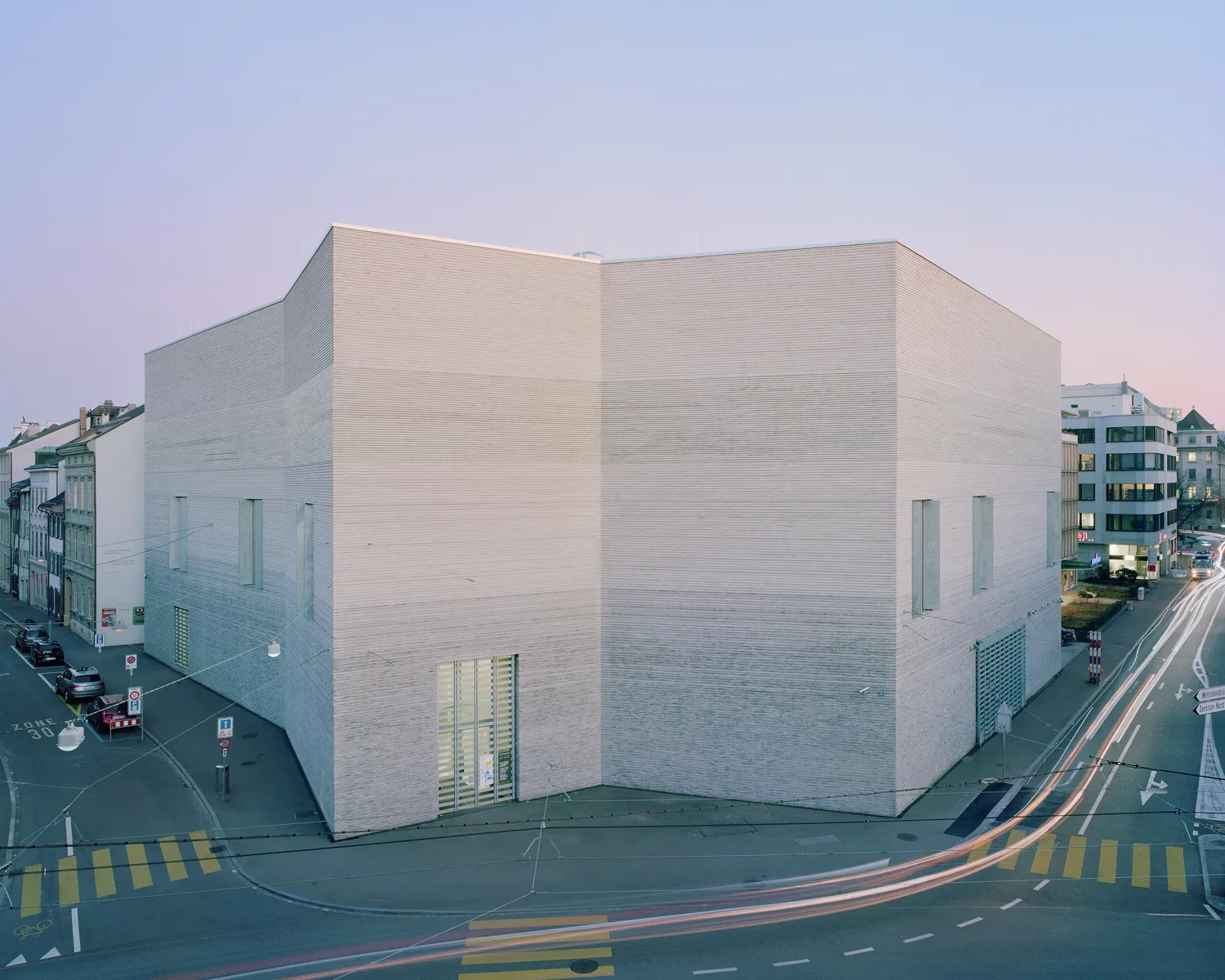 Groot hoekig wit kalkstenen gebouw met minimalistische geometrische gevel bij schemering, omringd door stadsstraten met lichtsporen van voorbijrijdend verkeer