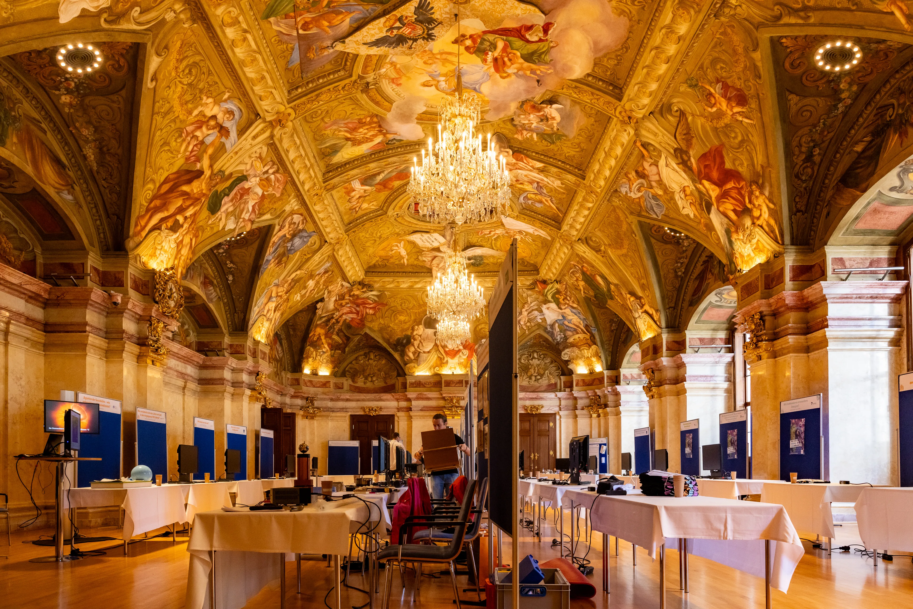 Ornamentowane wnętrze ze złoconymi freskami na suficie i żyrandolami, z wystawowymi stołami i panelami