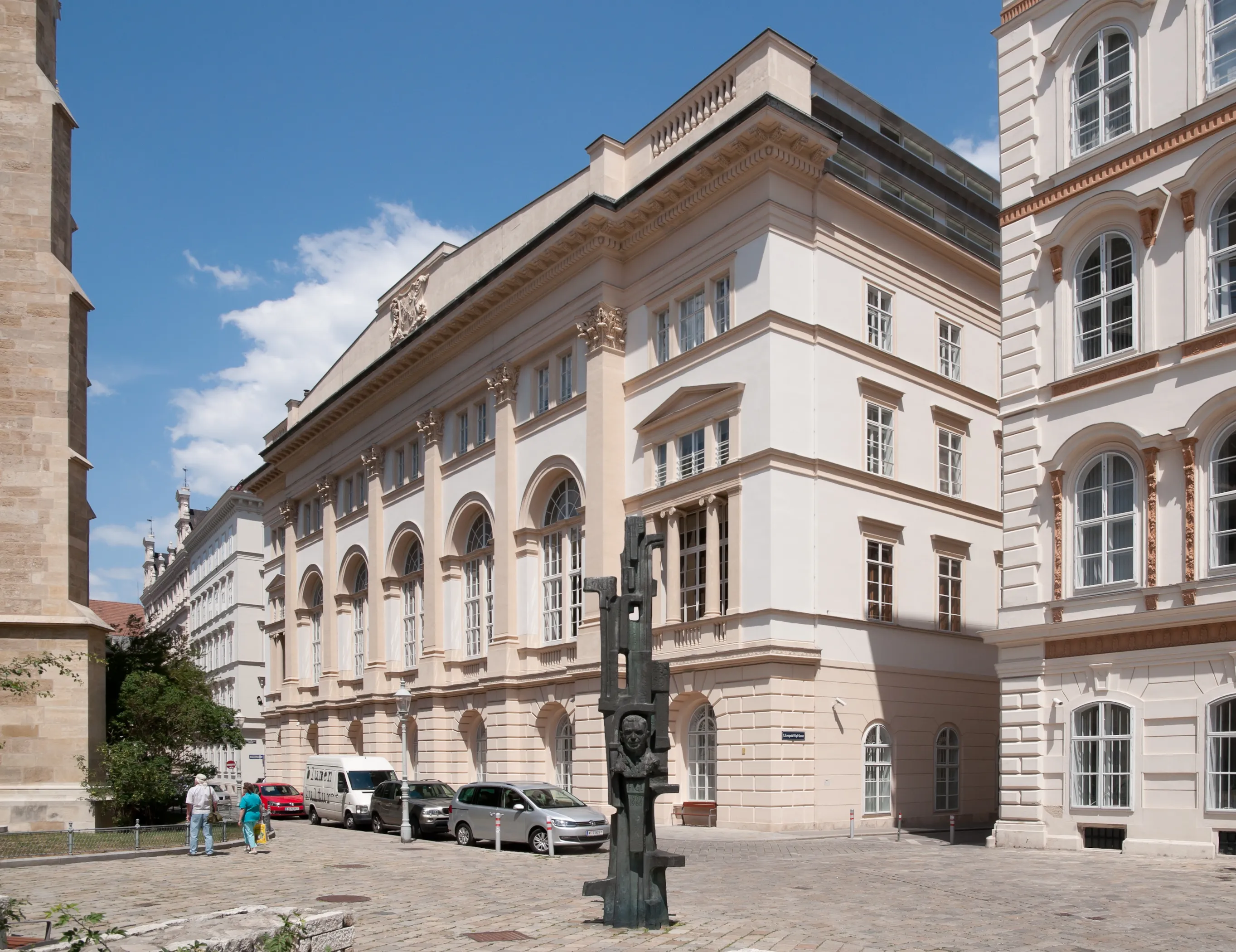 Duży, klasycystyczny budynek z łukowymi oknami i beżową fasadą, z nowoczesną rzeźbą na dziedzińcu.