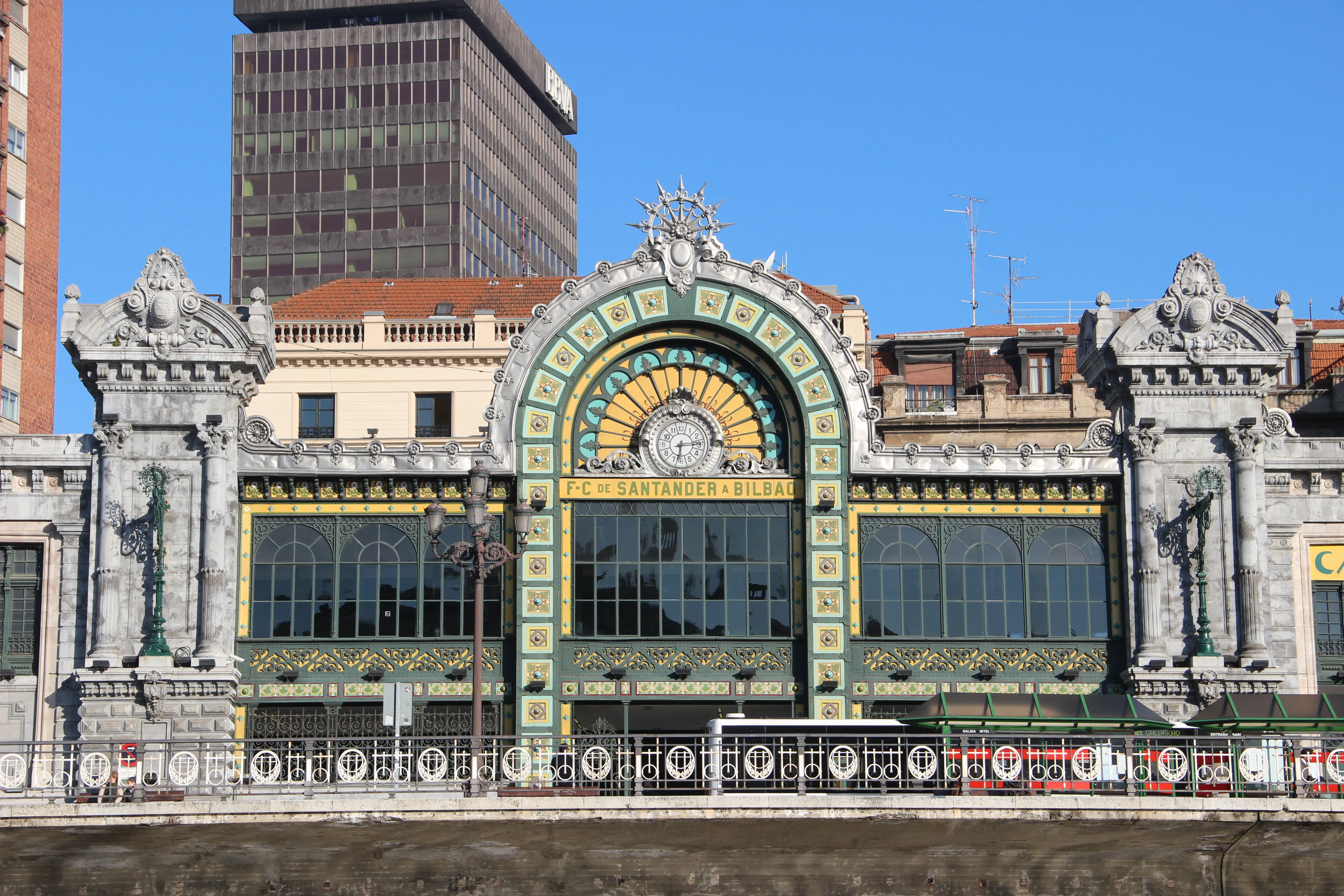 The ornate modernist façade of La Concordia Railway Station featuring a large arched central section with a clock, colorful ceramic tilework, ironwork windows, and decorative stone pillars on either side.