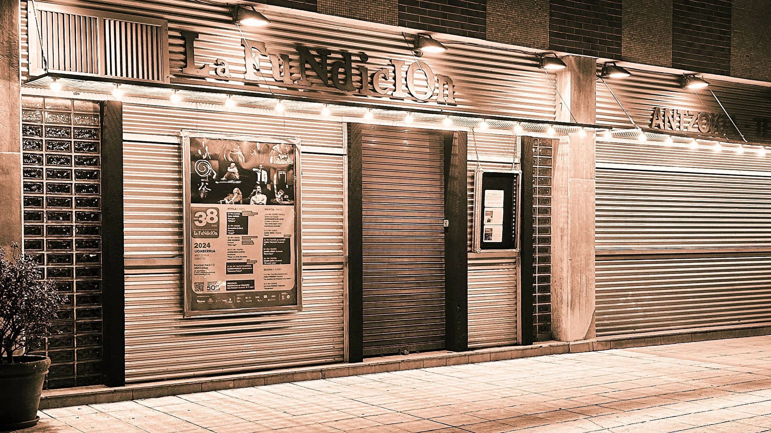 Nocny widok zewnętrzny na fasadę budynku La Fundición z zamkniętymi metalowymi roletami, podświetlanym szyldem i plakatami wydarzeń.