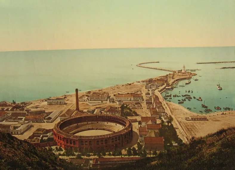 Ilustración histórica que muestra la plaza de toros circular Plaza de Toros de La Malagueta a lo largo de la costa de Málaga, con el puerto y el mar al fondo.