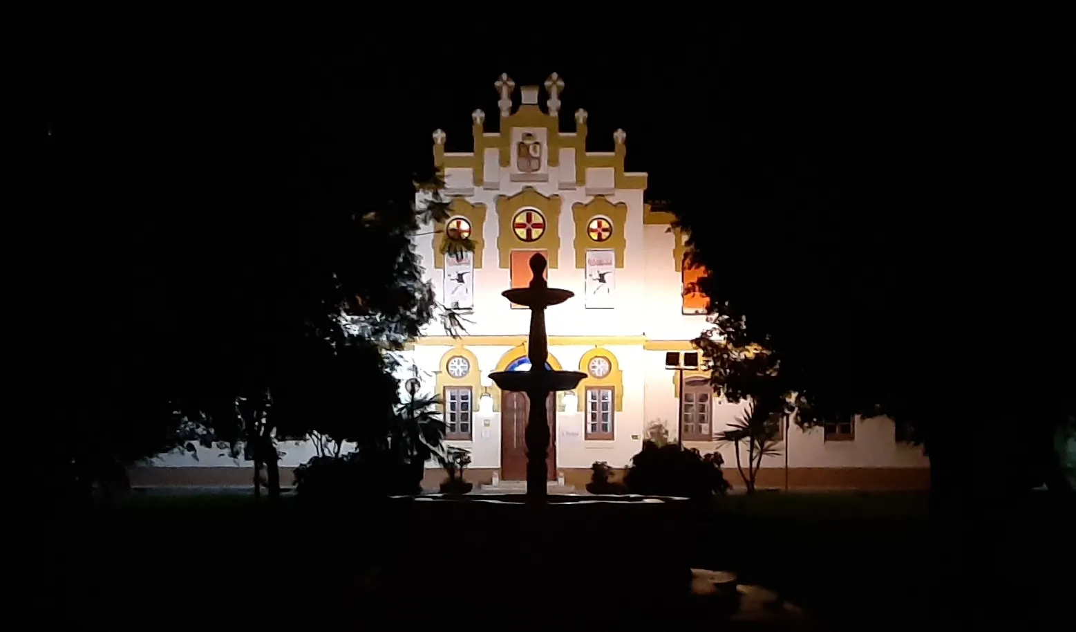 Vista nocturna de la fachada iluminada en blanco y amarillo de La Térmica con la silueta de una fuente en primer plano.