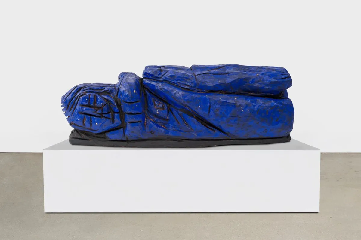 Una escultura grande de color azul brillante de una figura humana reclinada sobre un pedestal rectangular blanco en una galería.
