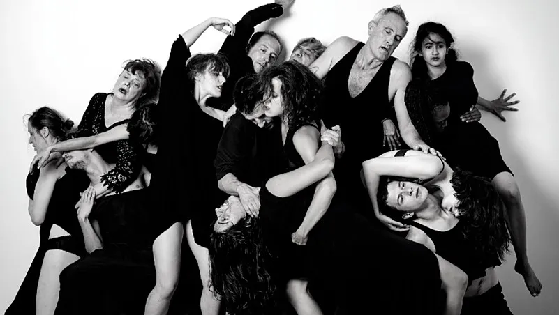 Grupo de artistas vestidos de negro posando dramáticamente en posiciones superpuestas sobre un fondo blanco, fotografía en blanco y negro