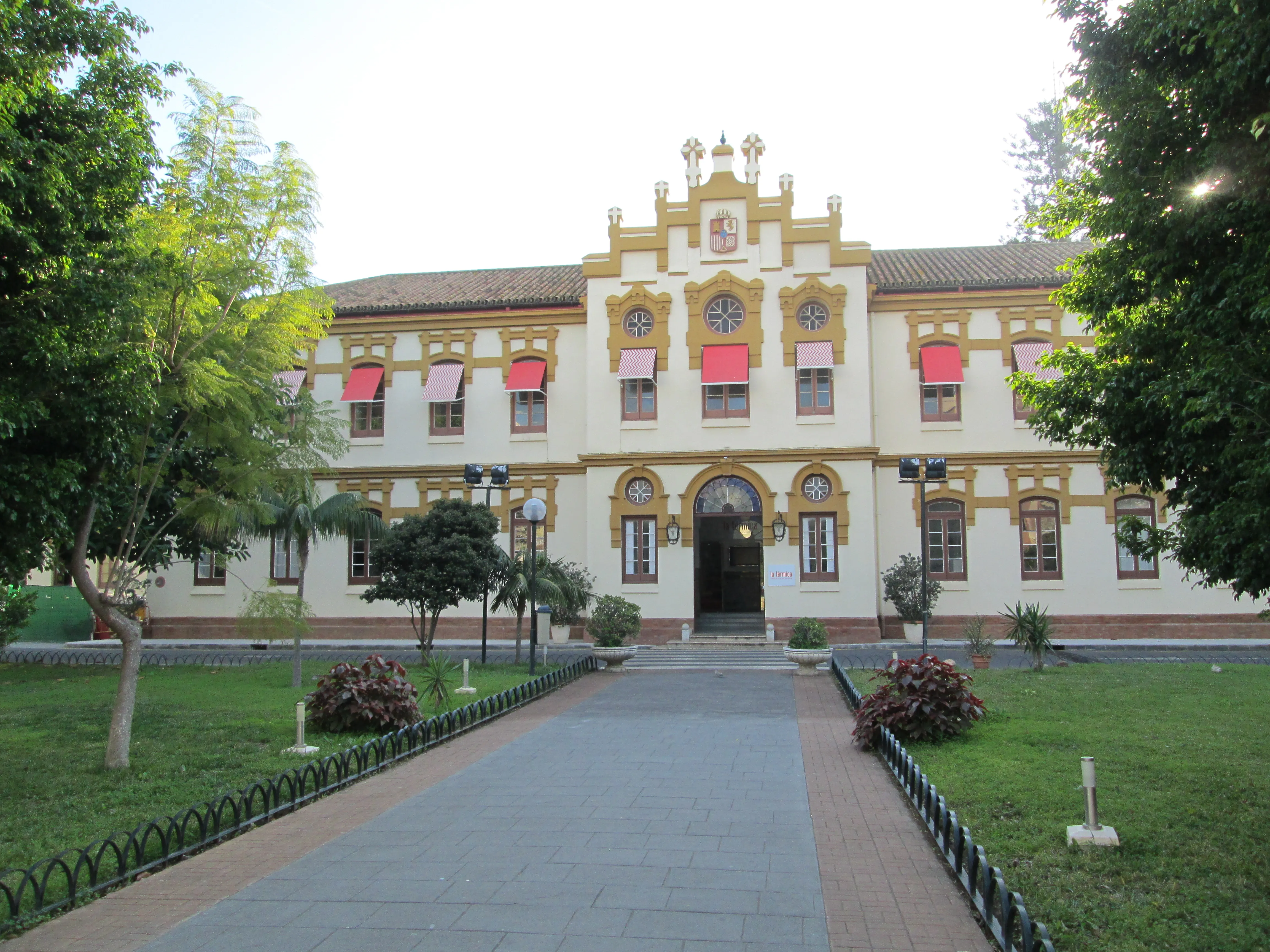 Fachada histórica blanca y amarilla con toldos rojos, entrada arqueada y un camino pavimentado que atraviesa un césped verde con árboles