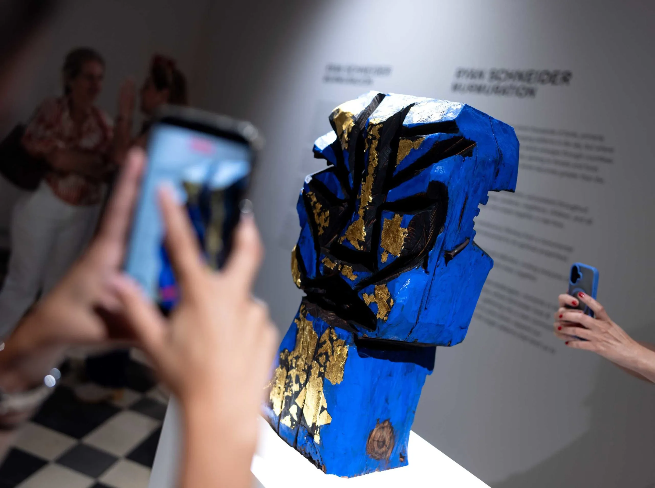 Escultura en azul, oro y negro sobre un pedestal blanco, fotografiada por visitantes con sus teléfonos.