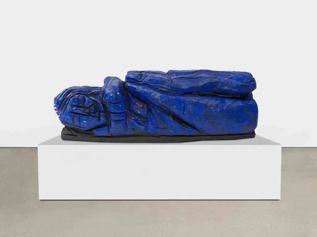 Una vibrante escultura azul de una figura humana reclinada sobre un pedestal rectangular blanco en una galería de paredes blancas.