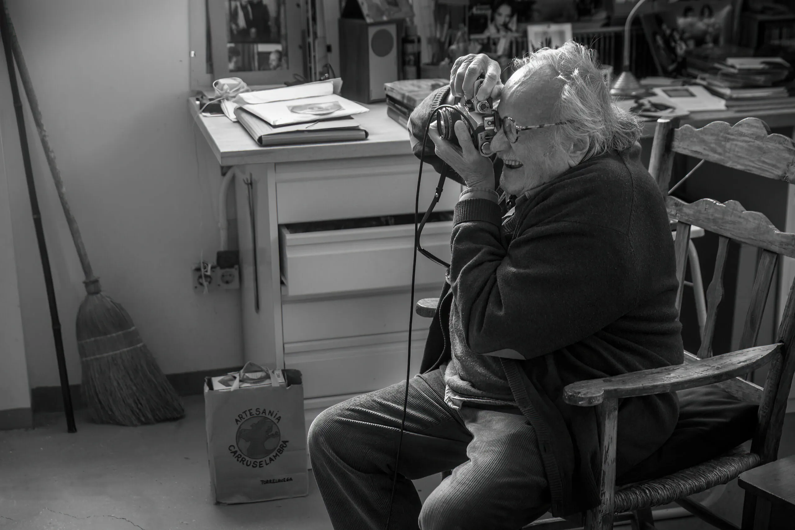 Fotografía en blanco y negro de un anciano con el pelo blanco y gafas sentado en una silla de madera, sosteniendo una cámara frente a su rostro en una habitación desordenada.