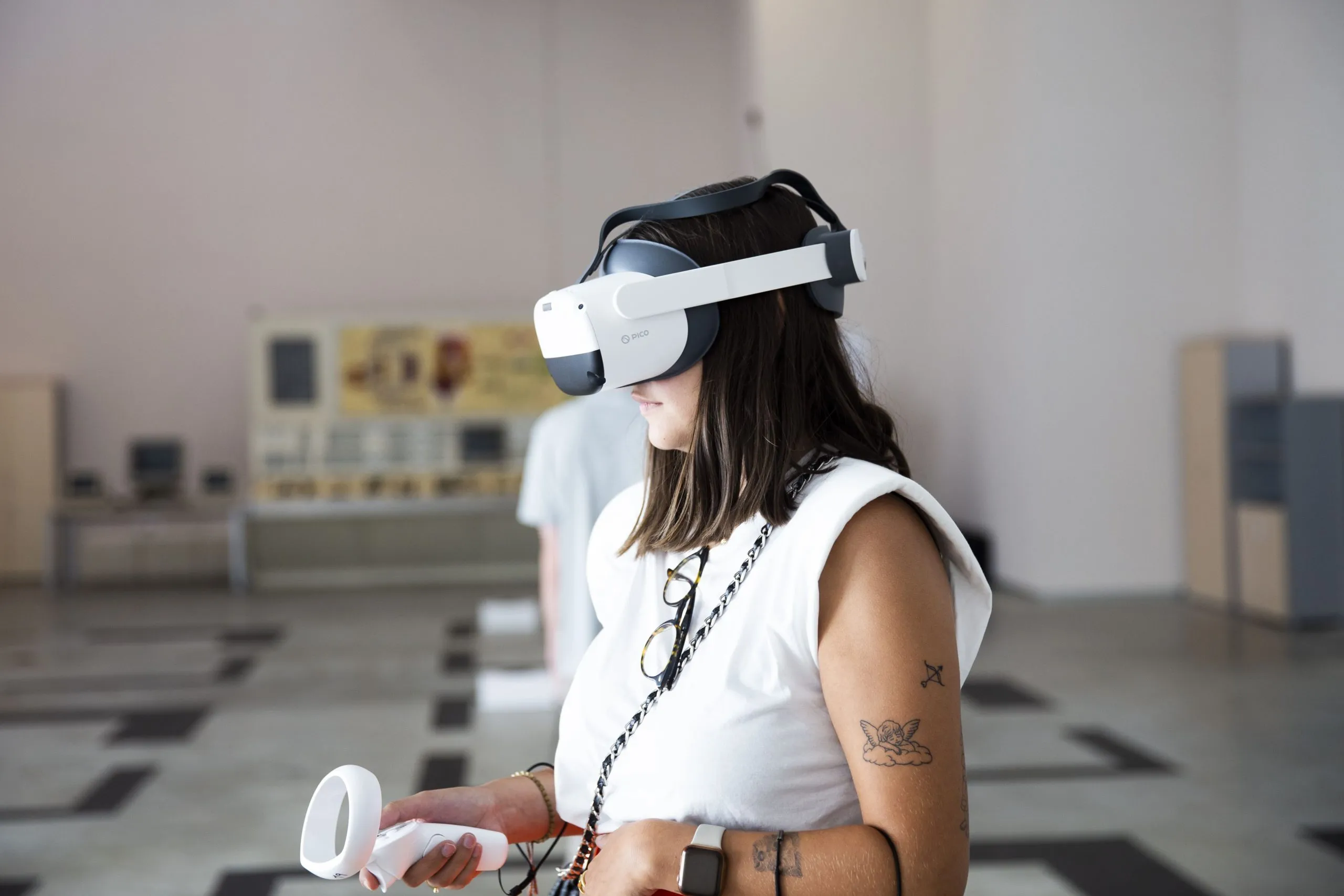 Una mujer que lleva un casco de realidad virtual blanco y sostiene un mando está de pie en una luminosa sala de exposiciones con paneles informativos al fondo.