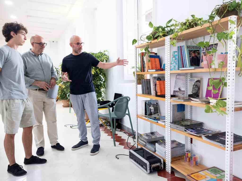Tres hombres están de pie en una habitación luminosa mirando unas estanterías de metal blanco llenas de libros, revistas y plantas colgantes, con uno de ellos señalando hacia los estantes.