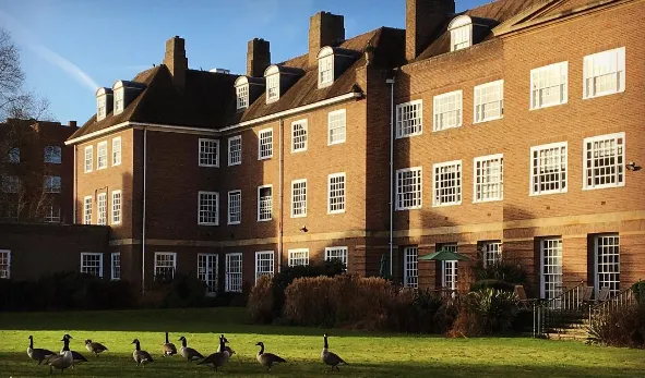 Rodbakstenen gebouw van meerdere verdiepingen met witomrande ramen en schoorstenen, met ganzen op een groen gazon op de voorgrond onder een blauwe lucht
