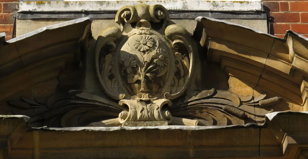 Close-up van een gebeeldhouwde stenen cartouche met florale motieven en krulwerk boven een boog, afgezet tegen rode baksteen.