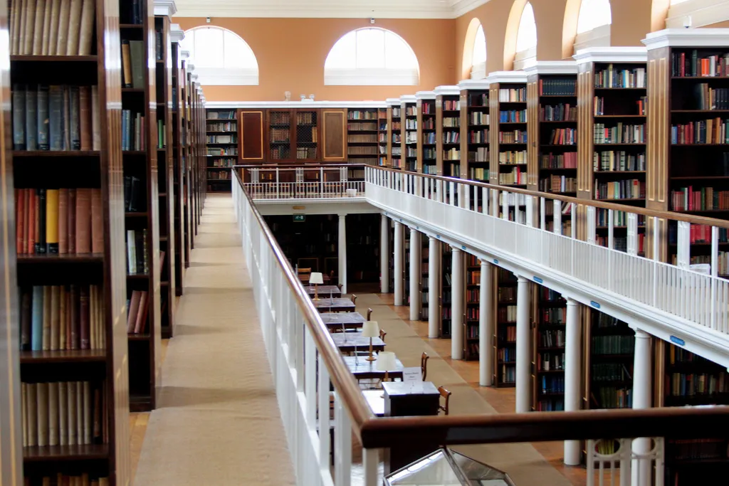 Dwupiętrowe wnętrze biblioteki z wysokimi drewnianymi regałami, antresolą z białą barierką, łukowatymi oknami oraz stolikami do czytania na dole.
