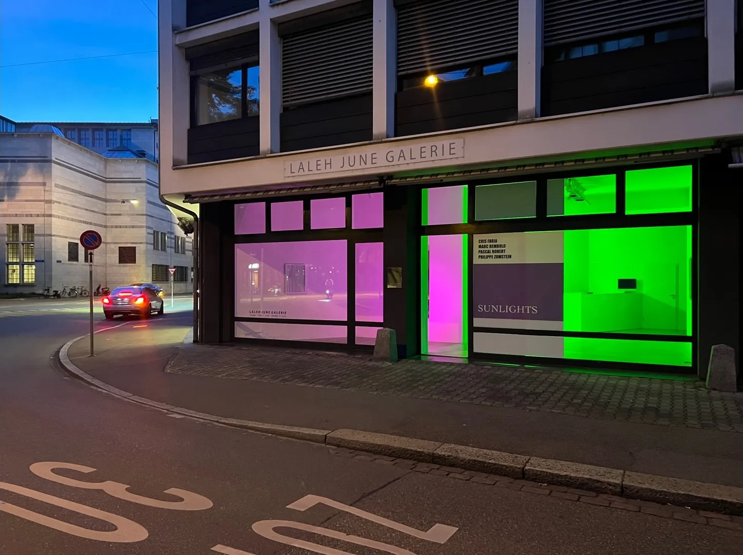 Vista exterior de Laleh June Galerie al atardecer con ventanas iluminadas en rosa y verde en una esquina