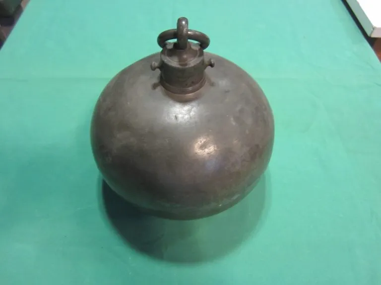 Un recipiente de metal vintage, redondo, con un asa en forma de anillo en la parte superior, sobre una superficie verde.