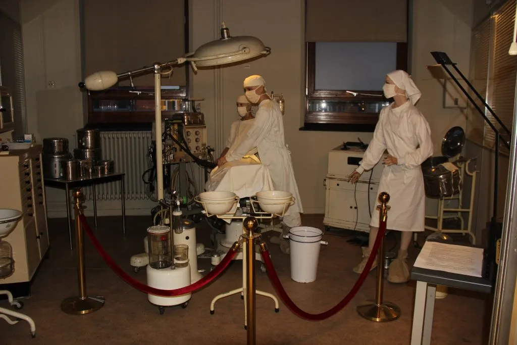 Exhibición de museo que muestra maniquíes vestidos como personal médico en un quirófano histórico, con una lámpara quirúrgica y equipo médico detrás de un cordón de terciopelo rojo.
