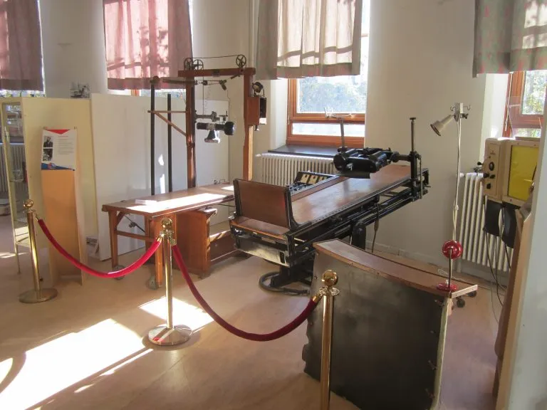 Mesa de operaciones médica antigua y equipo sanitario histórico expuestos tras cuerdas de terciopelo rojo en una sala de museo con grandes ventanales