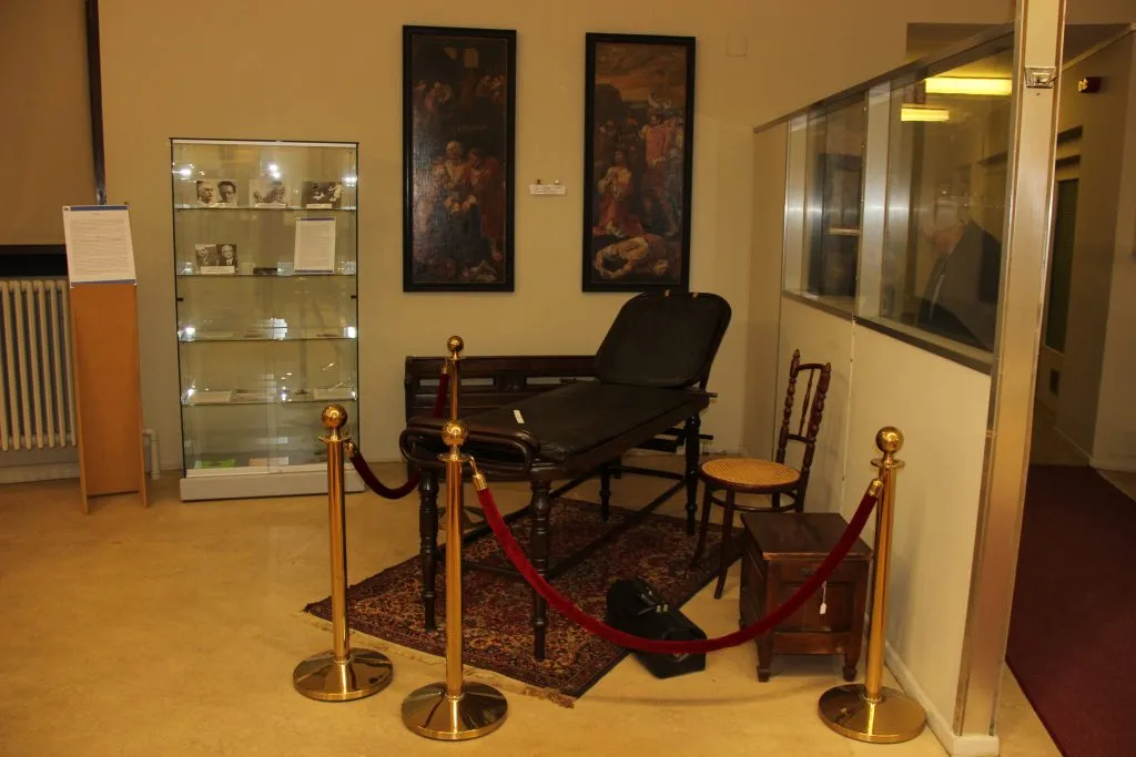 Interior de un museo que muestra una silla de reconocimiento médico histórica acordonada con postes de terciopelo rojo, flanqueada por una vitrina de cristal y dos cuadros enmarcados en la pared.