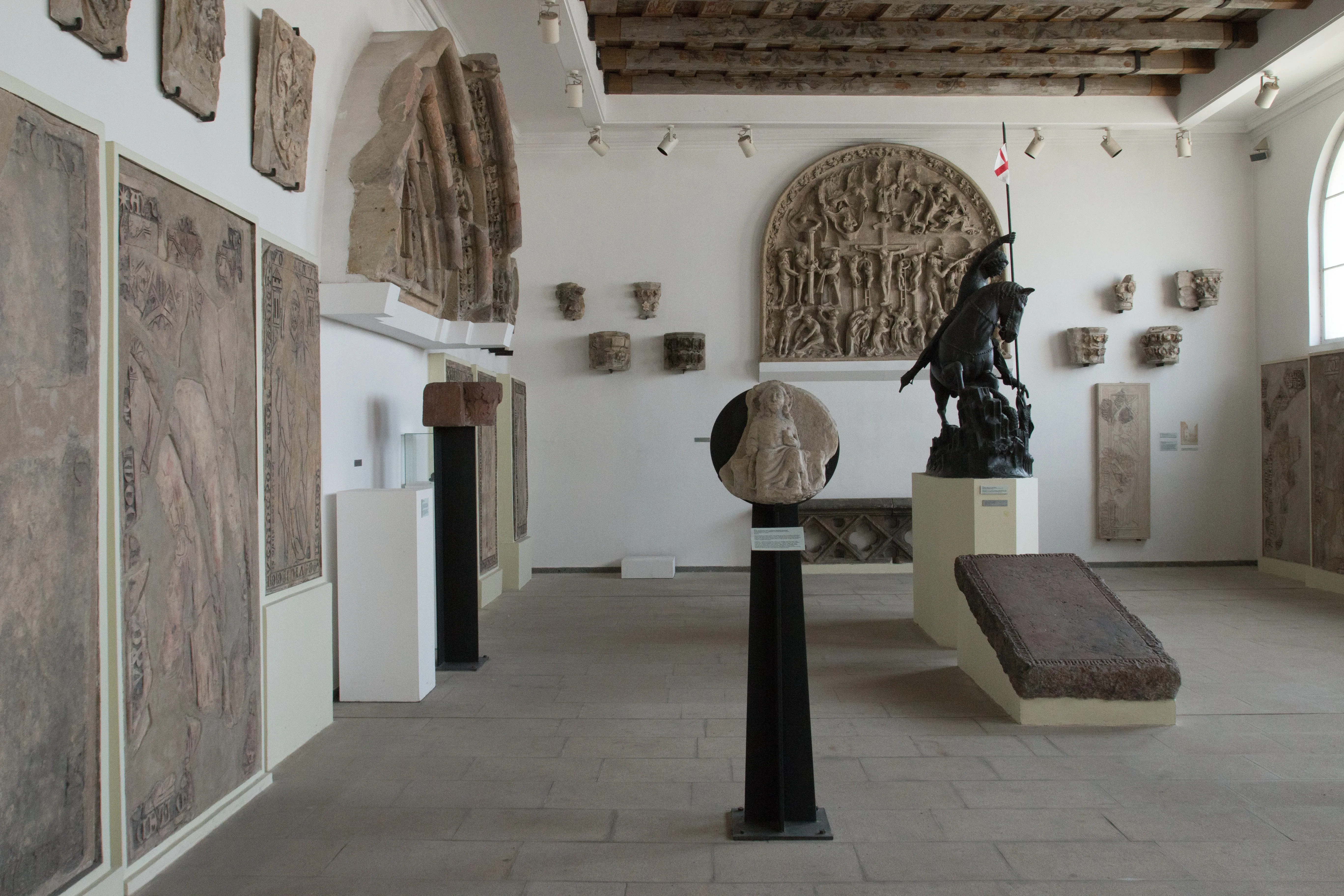 Interieur van een galerie met stenen sculpturen en architectonische fragmenten, waaronder een groot reliëf en een beeld op een sokkel.
