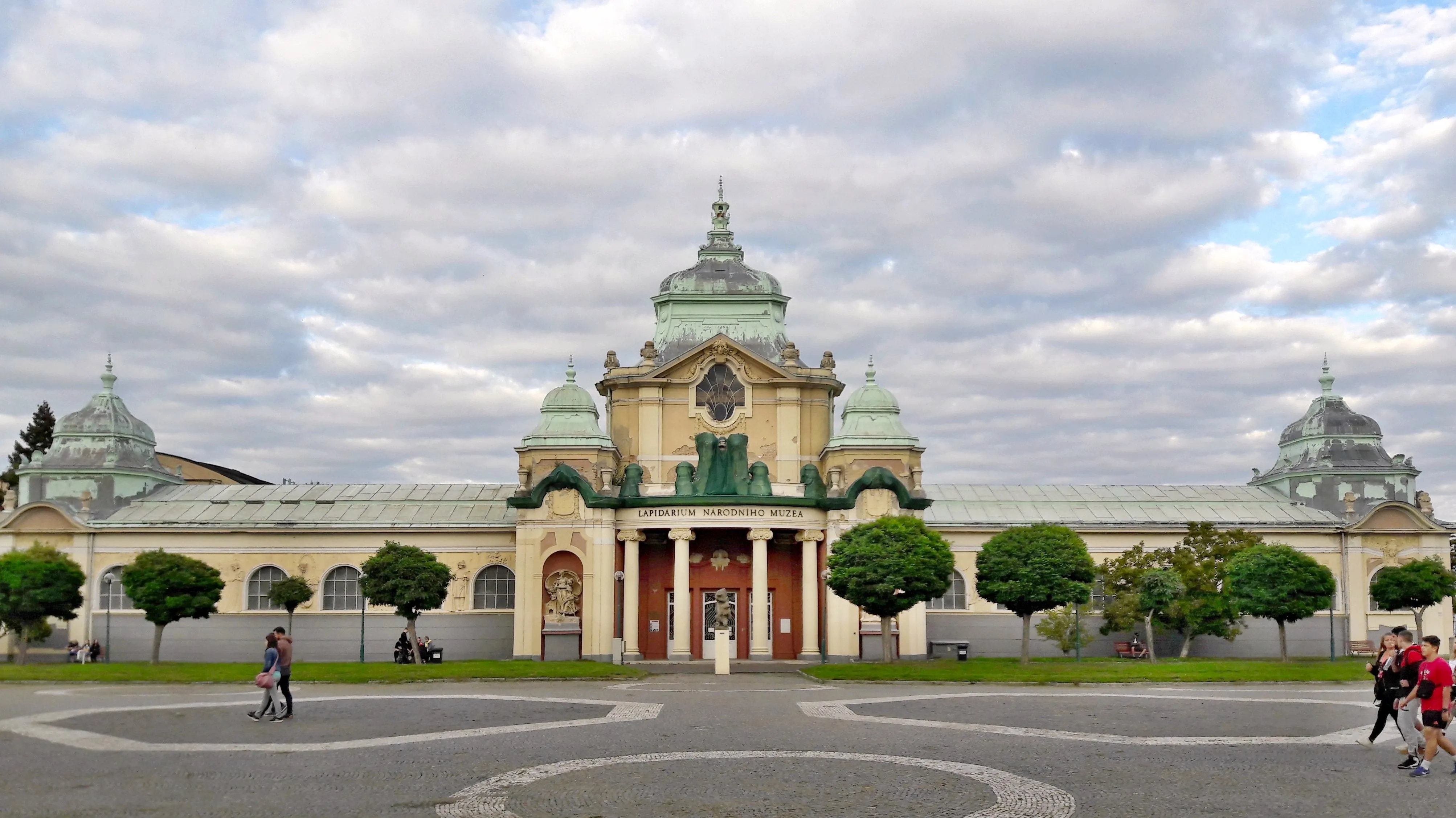 Een groot historisch gebouw met een centrale koepel en paviljoens met groene daken, omringd door getrimde bomen en een betegeld plein met wandelende mensen.