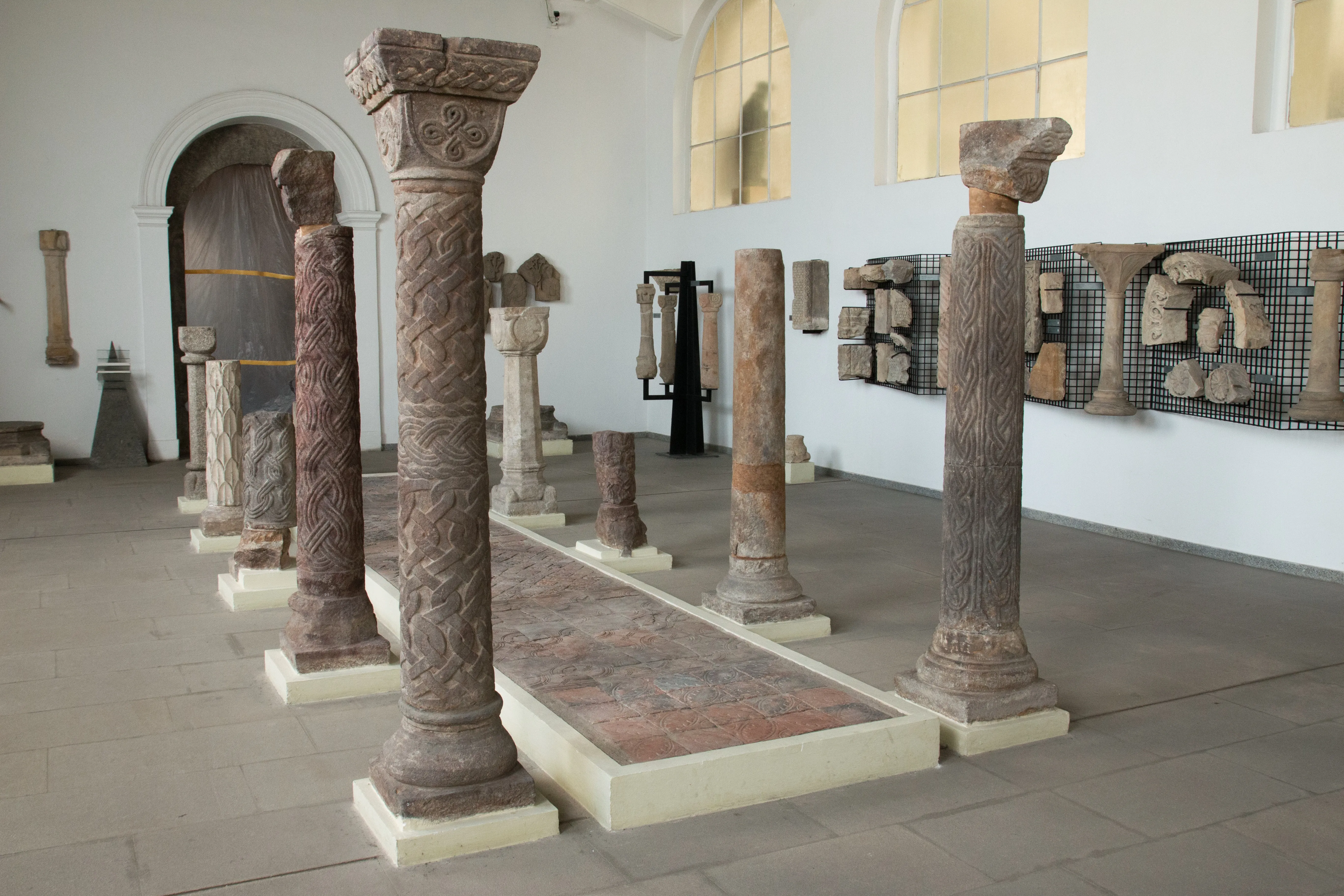 Een rij antieke stenen zuilen met gebeeldhouwde kapitelen in een museumgalerij, omringd door andere tentoongestelde stenen artefacten