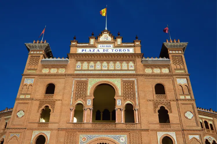 The facade of the Plaza de Toros de las Ventas with a sign reading 'PLAZA DE TOROS' and Spanish flags on top against a blue sky